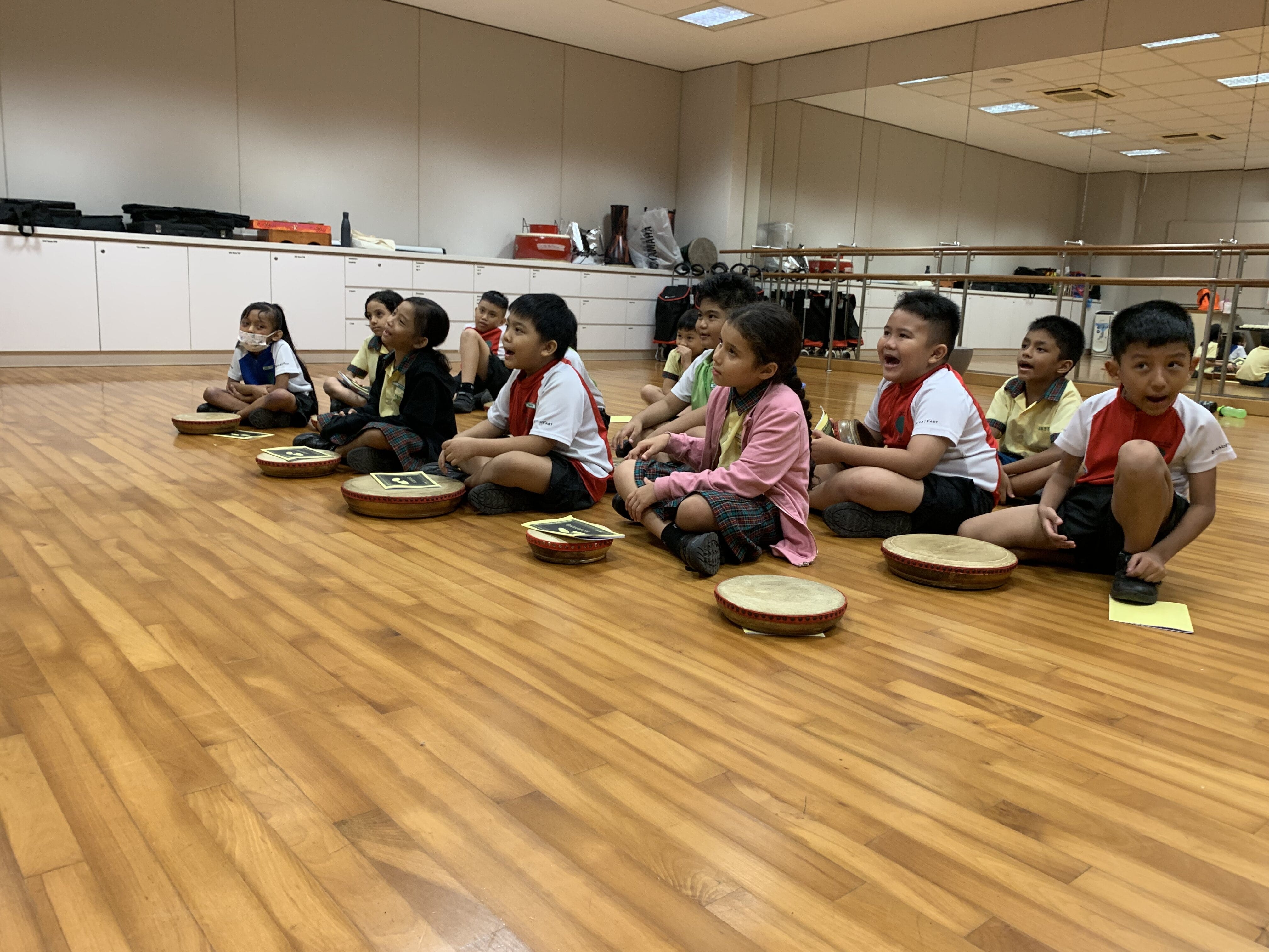Young children sit on a wood floor in a mirrored studio, each with a small hand drum, mouths open as if singing.